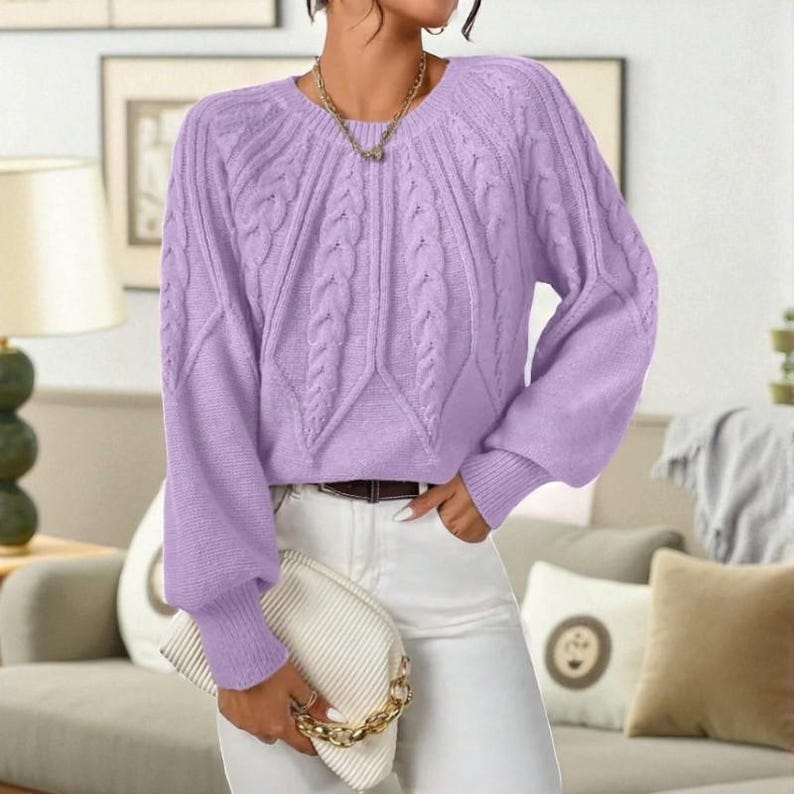May include: A light purple cable knit sweater with long sleeves. The sweater has a round neckline and a fitted waist. The sleeves are cuffed at the wrists. The person is wearing white pants and holding a white clutch with a gold chain.