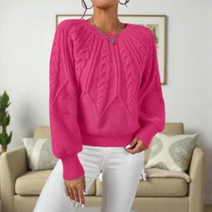 May include: A bright pink sweater with a cable knit design on the sleeves and front. The sweater has a round neckline and long sleeves with ribbed cuffs. The model is wearing white pants. The sweater is a vibrant, eye-catching piece.