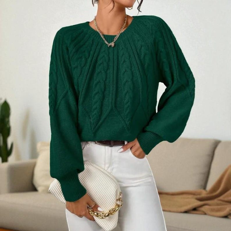 May include: A dark green cable knit sweater with long sleeves and a round neckline. The sweater has a textured design on the front and sleeves. The person is wearing white pants and holding a white clutch with a gold chain.