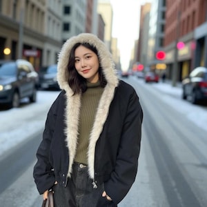 May include: A black hooded jacket with a cream-colored faux fur lining and hood. The jacket is worn over a green sweater and dark jeans. The background shows a city street with cars and snow.