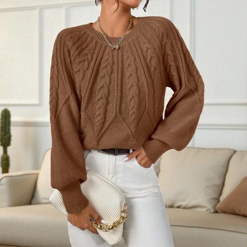 May include: A brown cable knit sweater with a round neckline and long sleeves. The sweater has a textured design on the front and sleeves. The person is wearing white pants and holding a white clutch with a gold chain.