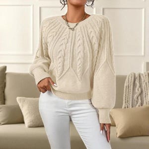 May include: Cream-colored cable knit sweater with a round neckline and long sleeves. The sweater features a textured cable knit design on the front and sleeves, paired with white pants. The model is wearing a gold necklace.