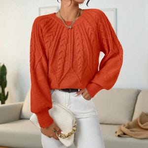 May include: An orange cable-knit sweater with long sleeves and a round neckline. The sweater has a textured design and ribbed cuffs. The person is wearing white pants and holding a white clutch with a gold chain.