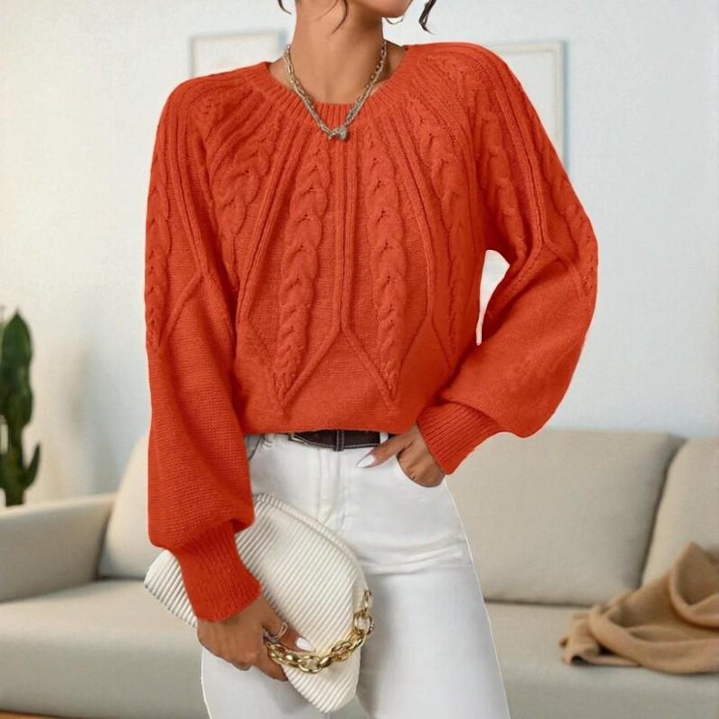 May include: An orange cable-knit sweater with long sleeves and a round neckline. The sweater has a textured design and ribbed cuffs. The person is wearing white pants and holding a white clutch with a gold chain.