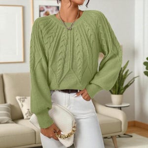 May include: A light green cable knit sweater with long sleeves and a round neckline. The sweater has a textured design on the front and sleeves. The person is wearing white pants and holding a white clutch with a gold chain.