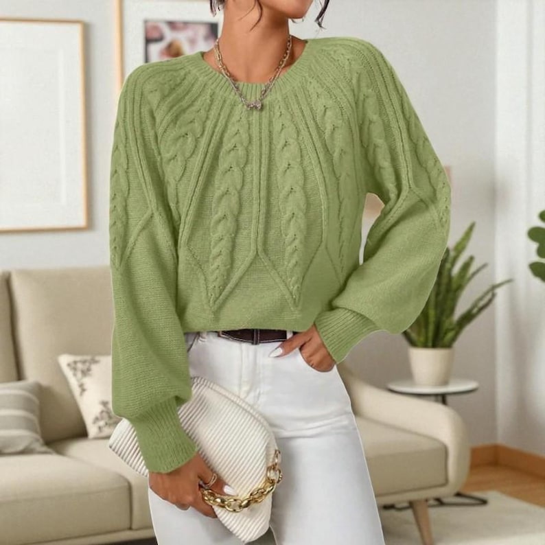 May include: A light green cable knit sweater with long sleeves and a round neckline. The sweater has a textured design on the front and sleeves. The person is wearing white pants and holding a white clutch with a gold chain.