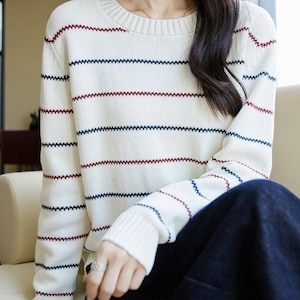 May include: Cream-coloured sweater with horizontal stripes in navy blue and burgundy. The sweater has a round neckline and long sleeves. The stripes are created with a zig-zag stitch. The sleeves end with a ribbed cuff.