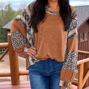 Op de afbeelding: Een bruine geribbelde top met lange mouwen, voorzien van patronen en luipaardprint. De top heeft een ronde halslijn en wordt gedragen met een blauwe jeans. De mouwen combineren verschillende patronen en dierenprints.