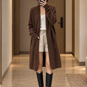 May include: A long, open-front brown knit cardigan with long sleeves and two front patch pockets. Underneath, a white ribbed tank top is paired with beige drawstring shorts. The outfit is completed with black knee-high boots.