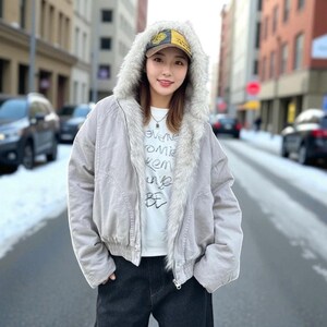 May include: A light gray jacket with a faux fur-lined hood and a matching baseball cap. The jacket has a zipper closure and is worn over a white t-shirt with text. The background shows a city street with cars and buildings.