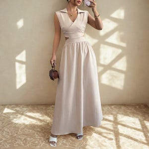 May include: A beige two-piece dress with a collared, sleeveless top and a long, flowing skirt. The top has a wrap-style design with a cutout at the waist. The dress is paired with white sandals and a small brown purse.