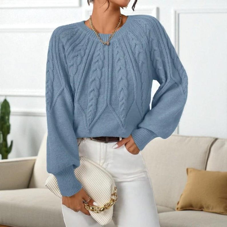 May include: A light blue cable knit sweater with long sleeves. The sweater has a round neckline and a textured cable knit design on the front and sleeves. The model is wearing white pants and holding a white clutch with a gold chain.
