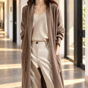 Könnte beinhalten: Eine lange, offene braune Strickjacke mit zwei aufgesetzten Vordertaschen, getragen über einem beigen V-Ausschnitt-Top und einer hellcremefarbenen Hose mit einem einzelnen dunklen Knopfverschluss am Bund.