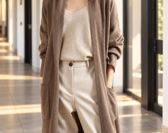 Könnte beinhalten: Eine lange, offene braune Strickjacke mit zwei aufgesetzten Vordertaschen, getragen über einem beigen V-Ausschnitt-Top und einer hellcremefarbenen Hose mit einem einzelnen dunklen Knopfverschluss am Bund.