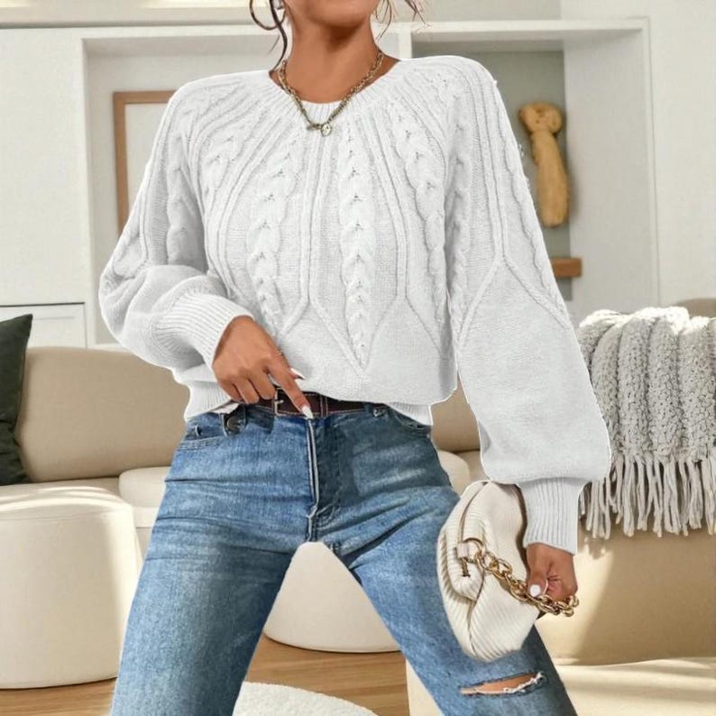 May include: A white cable-knit sweater with a round neckline and long, puffy sleeves. The sweater is paired with blue jeans and a small white purse with a gold chain strap. A gold necklace is also visible.