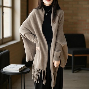 May include: A beige open-front knit cardigan featuring long sleeves with ribbed cuffs and a hemline adorned with thick, dangling fringe tassels. The garment is worn over a black high-neck top and black trousers, creating a layered appearance.