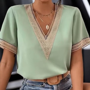 Op de afbeelding: Een lichtgroene blouse met een V-hals en korte mouwen. De halslijn en mouwranden zijn afgewerkt met een beige kanten design. De blouse wordt gedragen met een blauwe jeans en een bruine riem.