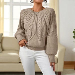 May include: A beige cable-knit jumper with a round neck and long sleeves. The jumper has a textured design with ribbed cuffs and hem. The model is wearing white trousers. The background includes a beige sofa and a window.