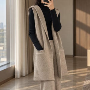 May include: A sleeveless beige knit waistcoat featuring a hood, an open front design, and two large patch pockets on the front. The garment is crafted with a ribbed texture along the edges and hem, providing a structured look over a black long-sleeve top.