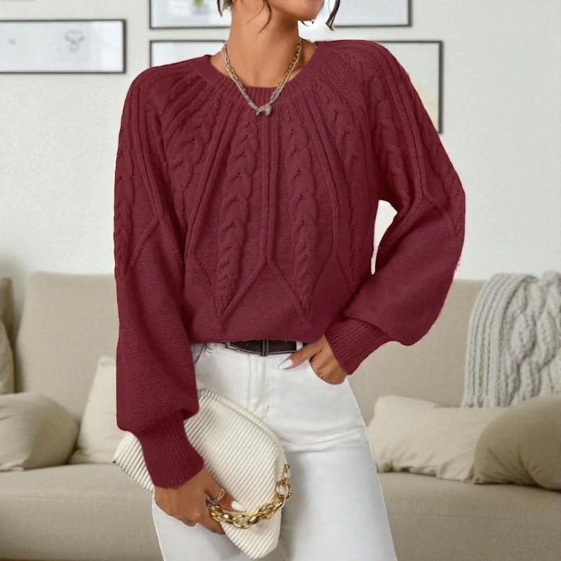May include: A burgundy cable-knit sweater with long sleeves and a round neckline. The sweater has a textured pattern on the front and sleeves. The sleeves have a ribbed cuff. The person is wearing white pants and holding a white clutch with a gold chain.
