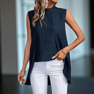 May include: Navy blue sleeveless top with a high neckline and a pleated front. The top has an asymmetrical hemline. The model is wearing white trousers and gold bracelets. This top is a stylish addition to any wardrobe.