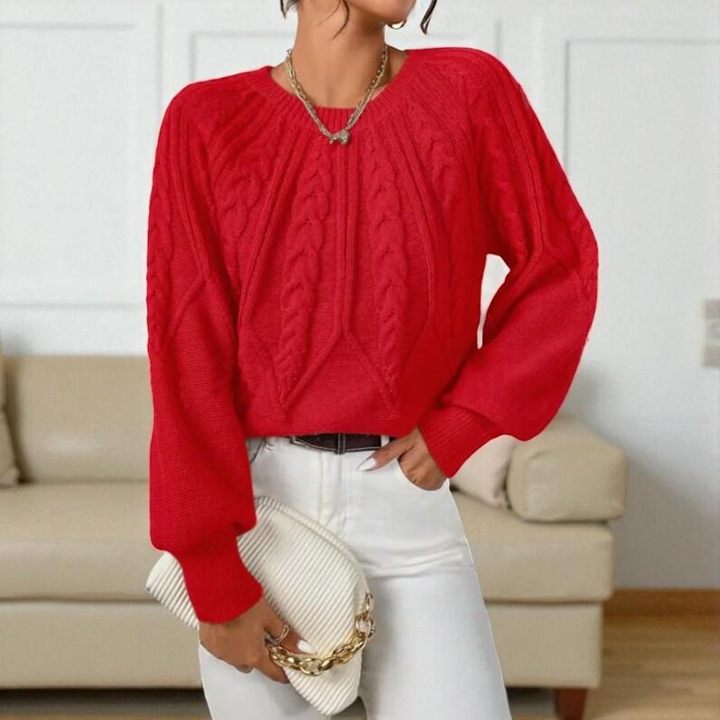 May include: A bright red cable-knit sweater with long sleeves. The sweater has a round neckline and a textured pattern. The person is wearing white pants, a gold necklace, and a white clutch with a gold chain.