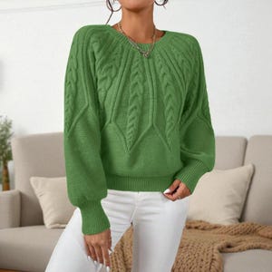 May include: A green cable-knit sweater with a round neckline and long sleeves. The sweater features a textured pattern on the front and sleeves. The cuffs and hem are ribbed. The model is wearing white pants.