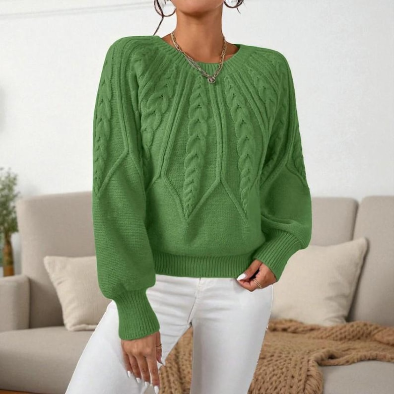 May include: A green cable-knit sweater with a round neckline and long sleeves. The sweater features a textured pattern on the front and sleeves. The cuffs and hem are ribbed. The model is wearing white pants.