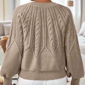 May include: A tan cable-knit sweater with a crew neck and long sleeves. The sweater features a textured pattern with vertical cable designs and a ribbed hem and cuffs. The sleeves have a slight balloon shape.