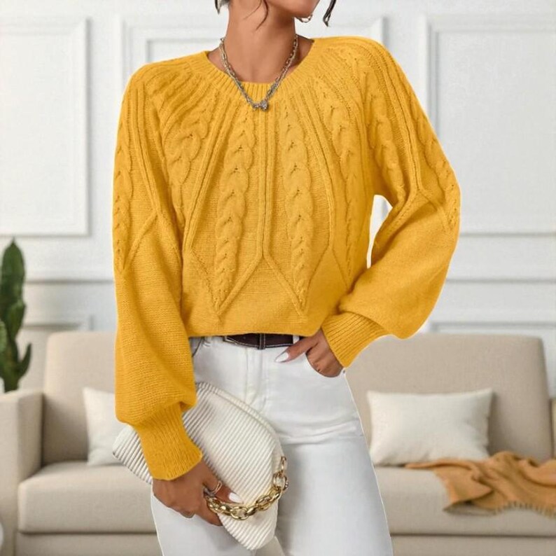 May include: A mustard yellow cable knit sweater with long sleeves and a ribbed cuff. The sweater has a V-neck and a textured cable knit design down the center. The model is wearing white pants and holding a white clutch.