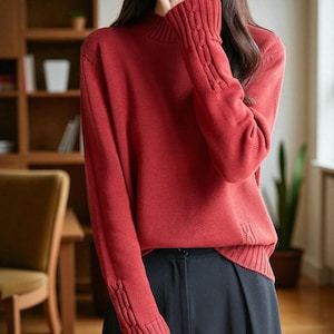 Può includere: Un maglione rosso a collo alto con polsini e orlo a coste. Le maniche presentano un motivo a maglia decorativo. Il maglione è abbinato a pantaloni neri. Lo sfondo comprende una libreria in legno e una sedia.