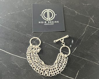 May include: Silver multi-chain bracelet with a toggle clasp. The bracelet features four chains of varying sizes and thicknesses. The bracelet is made by Noir Desire Jewellery.