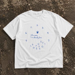 May include: A white short-sleeve t-shirt featuring a blue graphic of a clock face with numbers arranged in a circle, a small crown icon, and the handwritten text "Who cares I'm already late" with additional scattered numbers below the center.