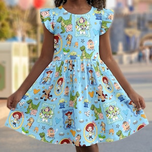 May include: A light blue dress featuring a pattern of cartoon characters. The dress has a round neckline and short, ruffled sleeves. The skirt flares out from the waist. The background is blurred, but appears to be a theme park.