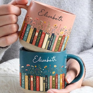 May include: A ceramic mug in dusty pink featuring the name Elizabeth and a row of colorful books with wildflowers. A second teal mug below it also displays the name Elizabeth and a similar row of books with wildflowers, both with speckled textures.