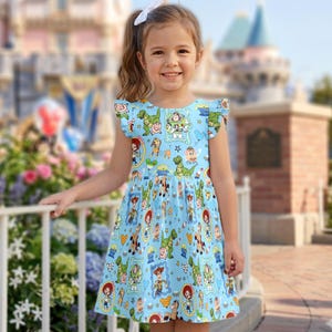 May include: A young girl wearing a light blue dress with a Toy Story character print. The dress has a round neckline and ruffled sleeves. The background shows a castle and flowers.