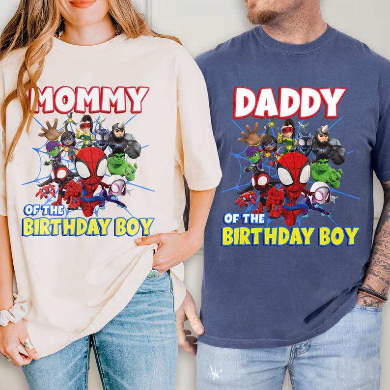 May include: A cream-colored t-shirt and a navy blue t-shirt, each featuring a graphic print of cartoon superheroes with a spiderweb background. The text reads MOMMY OF THE BIRTHDAY BOY or DADDY OF THE BIRTHDAY BOY in red and yellow lettering.