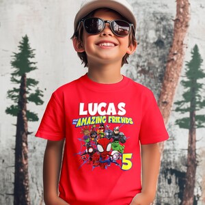 May include: A bright red short-sleeved t-shirt featuring a graphic print with the text LUCAS and his AMAZING FRIENDS, a group of cartoon superhero characters, blue spider web patterns, and a large yellow number 5 on the lower right side.