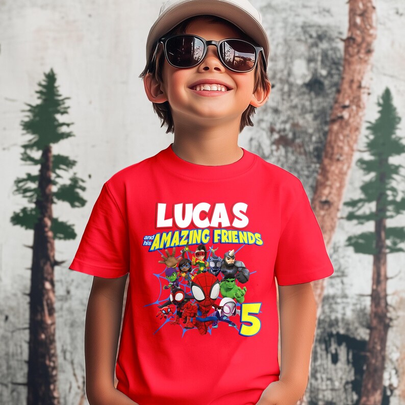 May include: A bright red short-sleeved t-shirt featuring a graphic print with the text LUCAS and his AMAZING FRIENDS, a group of cartoon superhero characters, blue spider web patterns, and a large yellow number 5 on the lower right side.