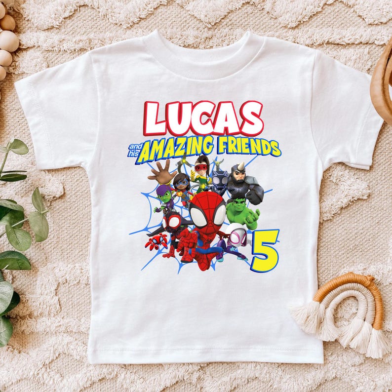 May include: A white short-sleeved t-shirt featuring a graphic print with the text LUCAS and his AMAZING FRIENDS. The design includes a group of cartoon superhero characters, a blue spiderweb background, and a large yellow number 5 on the right.