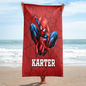 May include: Red beach towel featuring Spider-Man in a dynamic pose, set against a spiderweb design. The name "KARTER" is displayed in white block letters. The towel is held up on a beach with the ocean and sky in the background.