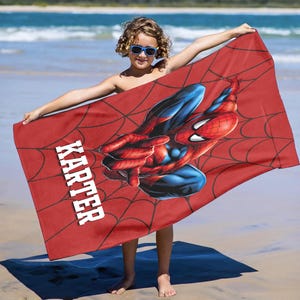 May include: Red beach towel featuring a Spider-Man graphic with a web design. The towel has the name "KARTER" printed in white. The towel is being held up by a person on a beach.