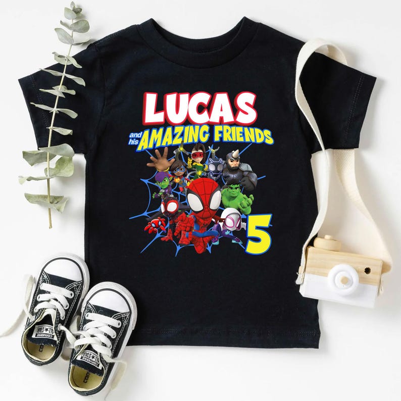 May include: A black t-shirt featuring a graphic print with the text LUCAS and his AMAZING FRIENDS, accompanied by a group of cartoon superhero characters, a blue spiderweb design, and a large yellow number 5 on the front of the garment.