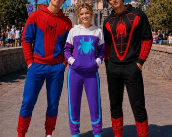 May include: Three people wearing matching hooded sweatshirts and sweatpants. The left outfit is red and blue with a black spider emblem. The center outfit is white, purple, and teal with a spider graphic. The right outfit is black and red with a spider web design.