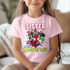 May include: A light pink short-sleeved t-shirt featuring a graphic print of various superhero characters and a blue spiderweb design. The text SISTER OF THE BIRTHDAY BOY is printed in red and yellow lettering above and below the character illustration.
