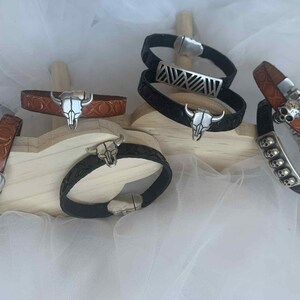 Op de afbeelding: Een verzameling leren armbanden met zilverkleurige versieringen. De armbanden hebben ontwerpen zoals een stierschedel, een schedel en geometrische patronen. De leerkleuren zijn bruin en zwart.