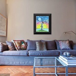 May include: A framed painting of a colorful tree with a person standing in the middle of it. The tree is surrounded by a blue sky with a moon and a yellow sunset. The painting is in a black frame and is hanging on a wall in a living room.