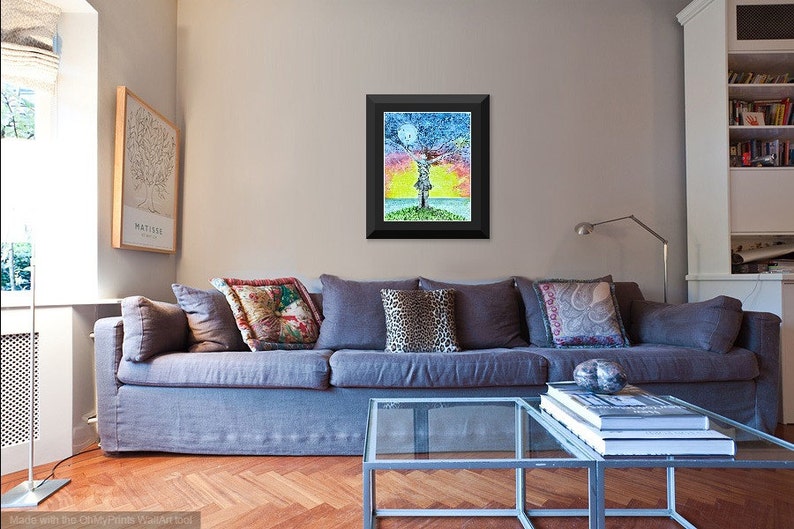 May include: A framed painting of a colorful tree with a person standing in the middle of it. The tree is surrounded by a blue sky with a moon and a yellow sunset. The painting is in a black frame and is hanging on a wall in a living room.