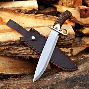May include: A large hunting knife with a polished steel blade and a handle made of deer antler. The knife has a brass bolster and a finger guard. It is displayed with a dark brown leather sheath with decorative stitching.