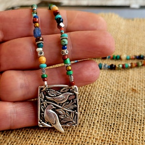 May include: A handcrafted necklace featuring a silver pendant with two birds perched on branches. The necklace has a colorful beaded strand with turquoise, red, brown, and blue beads. The pendant is rectangular with a detailed design.